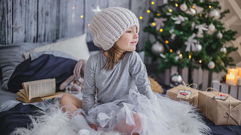 Cute Little Girl With Woolen Knitted Cap Is Wearing Ash Dress Sitting On Bed In Christmas Tree Background Cute, HD wallpaper