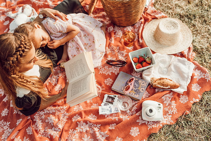 woman reading with girl while lying on orange and white floral picnic mat, HD wallpaper