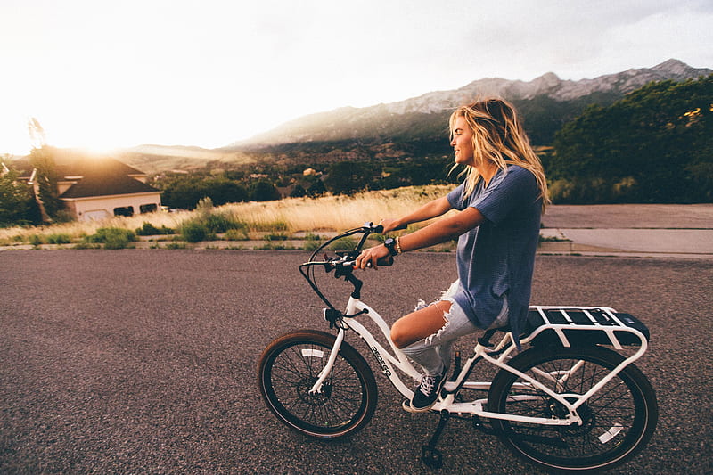 woman in blue t-shirt and distressed blue denim jeans riding white bicycle on grey road near mountains at daytime, HD wallpaper