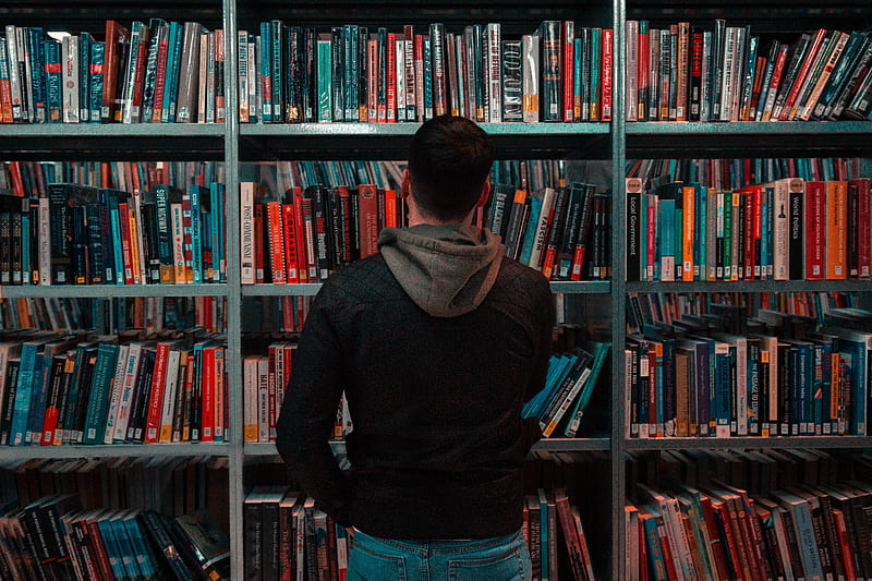 person wearing black and gray jacket in front of bookshelf, HD wallpaper