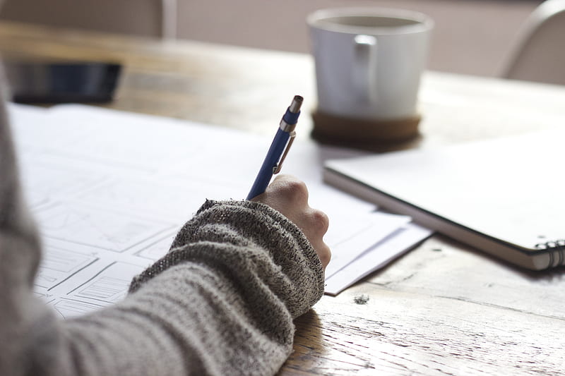 person writing on brown wooden table near white ceramic mug, HD wallpaper