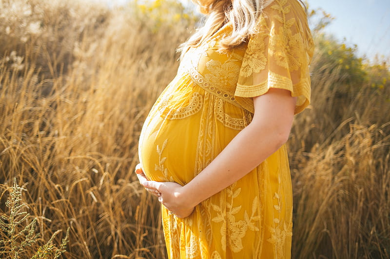 pregnant woman wearing yellow floral dress standing while touching her tummy and facing her right side near brown field during daytime, HD wallpaper