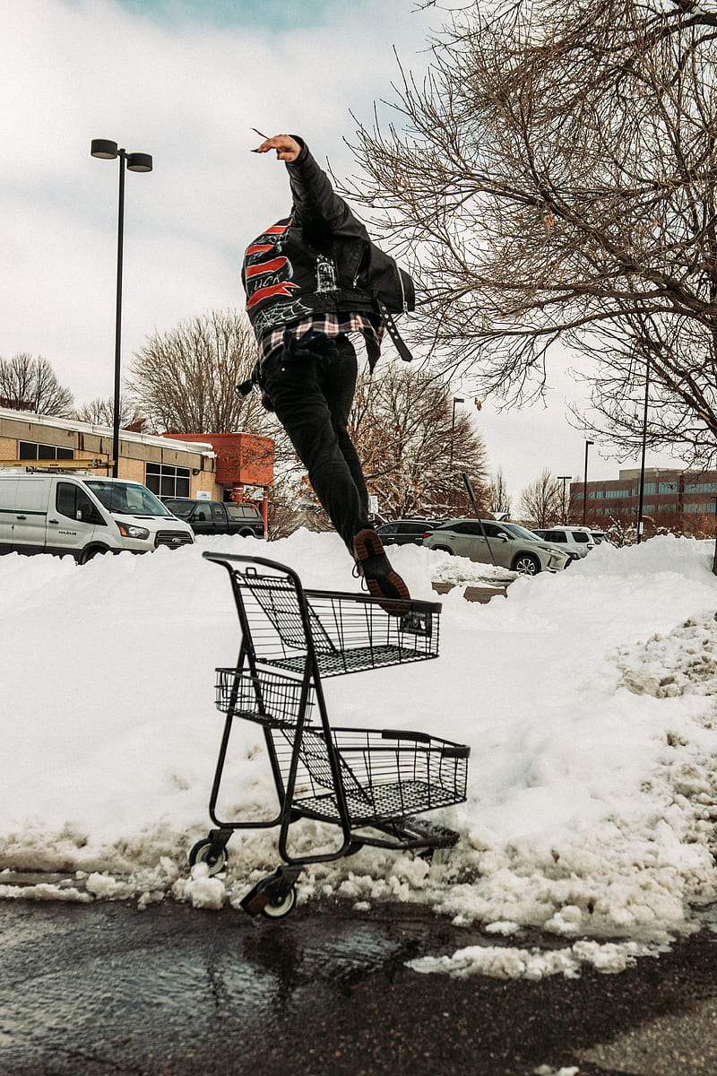man in black jacket and brown pants riding on black shopping cart on snow covered ground, HD phone wallpaper