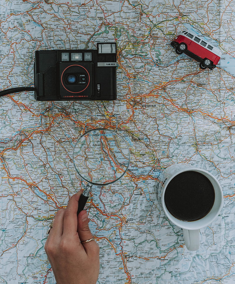 person holding magnifying glass beside black film camera white ceramic mug filled with black liquid near vehicle toy scale model, HD phone wallpaper