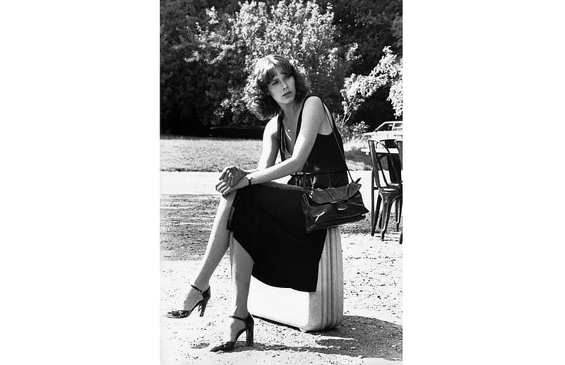 Sylvia Kristel, brunette, sitting on suitcase, flowers and bushes, black dress, spaghetti straps, hand bag, heels, jewelry, long strap, HD wallpaper