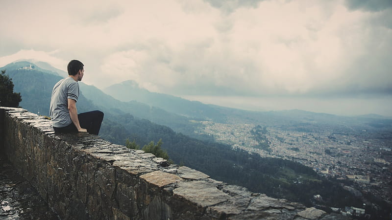 man sitting alone on concrete brick wall facing mountain and city under cloudy sky, HD wallpaper