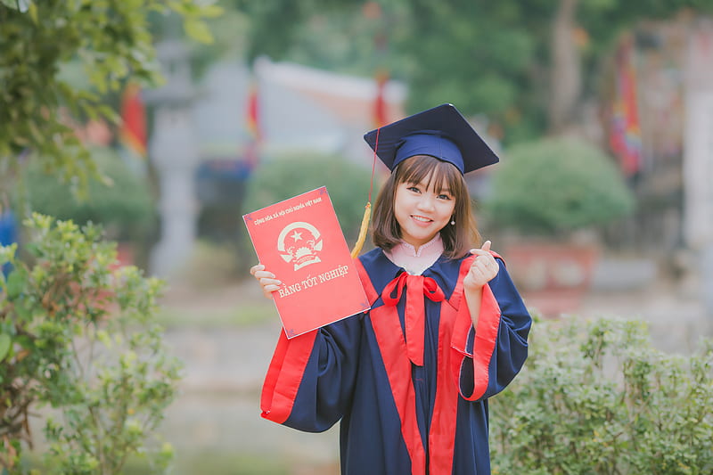 woman wearing black and red academic dress and mortar board holding red book cover near green grass, HD wallpaper