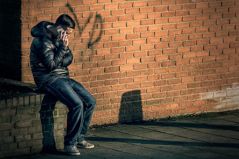 man in black leather jacket sitting on brick wall holding smartphone, HD wallpaper