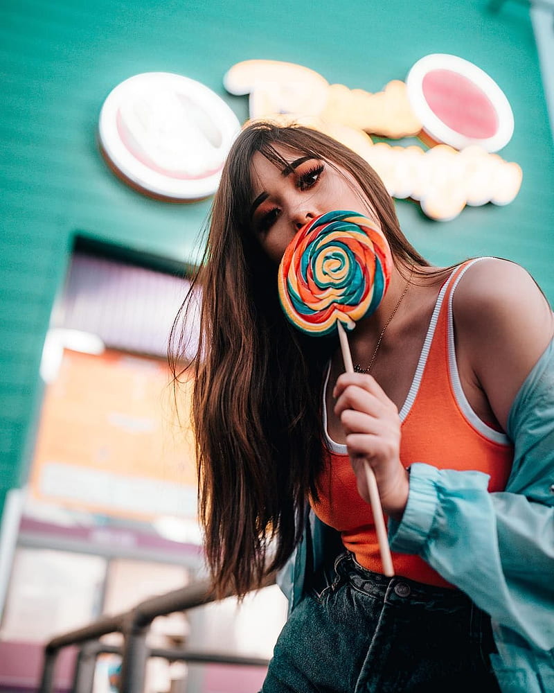 women, model, brunette, long hair, straight hair, eyeshadow, lollipop, jacket, women outdoors, vertical, bokeh, natural light, looking at viewer, tank top, jeans, orange clothing, leaning, hair in face, arched back, necklace, colorful, green, HD phone wallpaper