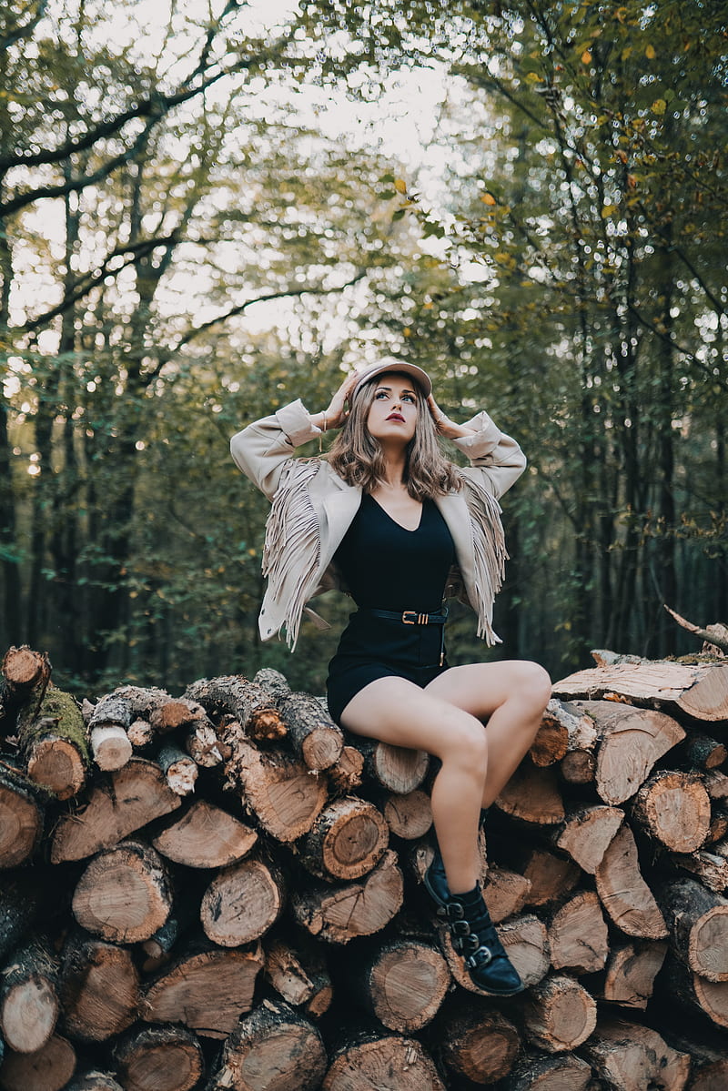 woman sitting on firewood stack looking up while touching her hat surrounded with green trees, HD phone wallpaper