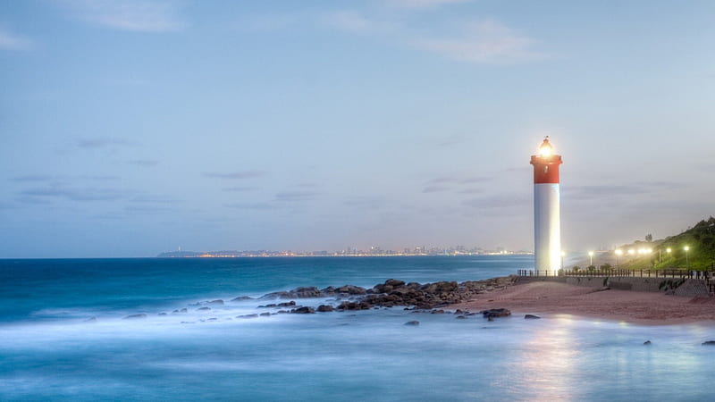 marvelous lit lighthouse with city in the background, city, seashore, dusk, lighthouse, lights, HD wallpaper